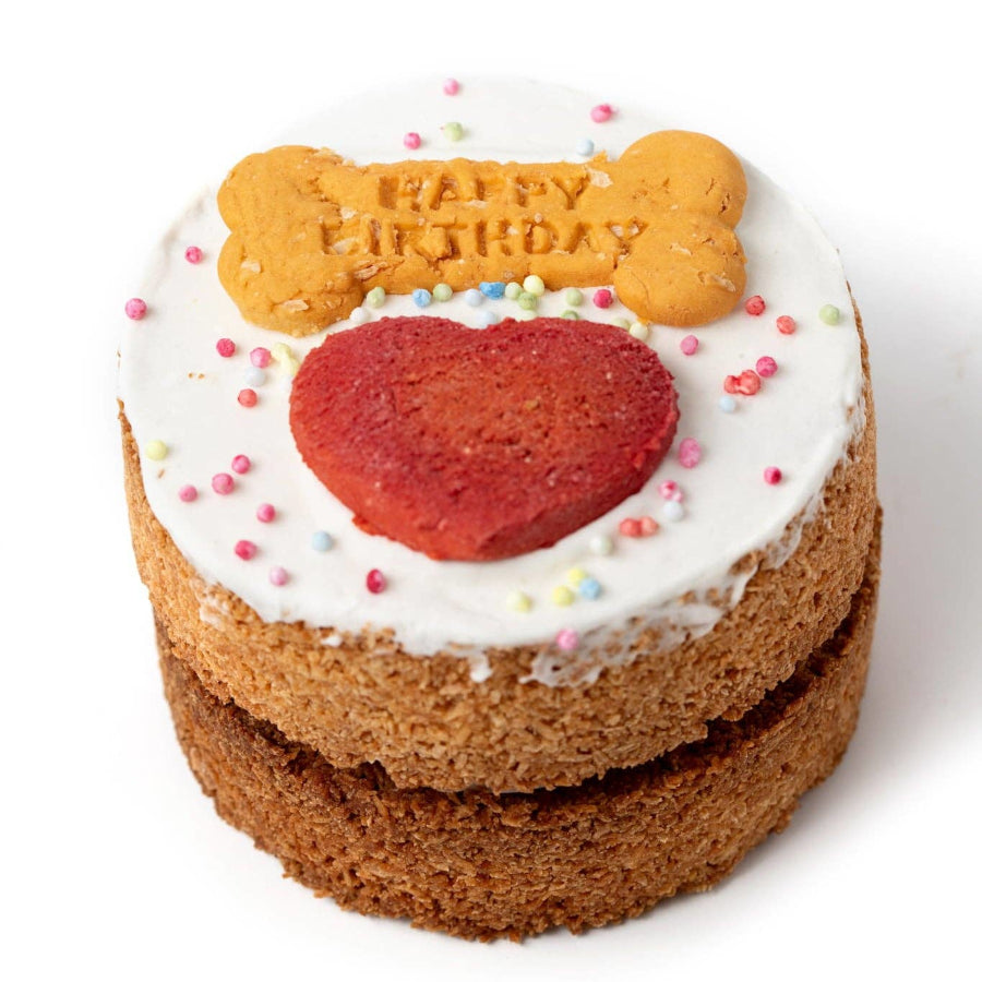 A two-layered cake decorated with white frosting, a red heart-shaped cookie, a golden-brown bone-shaped cookie with 'Happy Birthday' written on it, and colorful sprinkles, set against a plain white background.