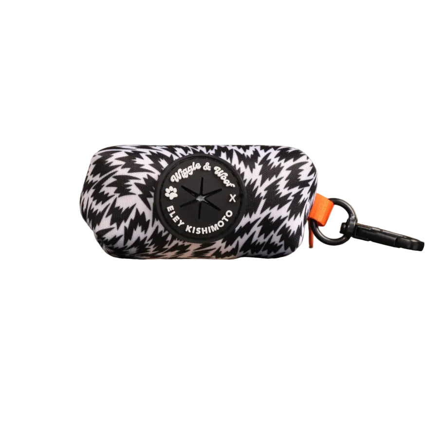 A cylindrical poo bag holder with a black and white flash pattern, featuring a central black circular patch with text. It has a black carabiner attached with an orange loop accent